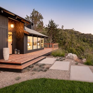 Designer concrete slabs lead the pathway to an expansive wood deck surrounding a rustic ebony black cabin perched upon a hill overlooking a lush California Canyon.