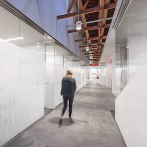 A woman walks down a brightly lit corridor flanked by a glass feature wall under wood trusses in a creative office environment