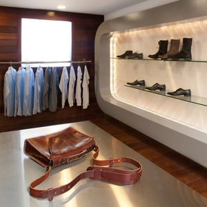 A retail store interior with a leather bag placed on a steel table in the foreground and a rack of dress shirts and shelves of shoes in the background.