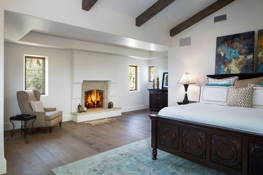 Exposed ceiling beams and a stone fireplace define this serene master suite’s traditional home design inspired by Spanish Colonial Revival architecture.