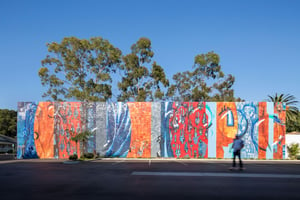 A skater boarder passes along a large public art mural spanning the length of a parking lot in fornt of a blue sky and tree line
