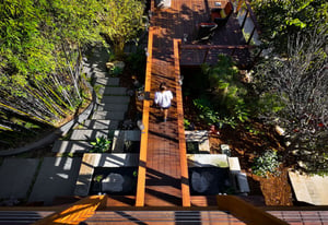 A woman walks along a suspended wooden ramp overlooking a tropical asian landscape and modern stepping patio.