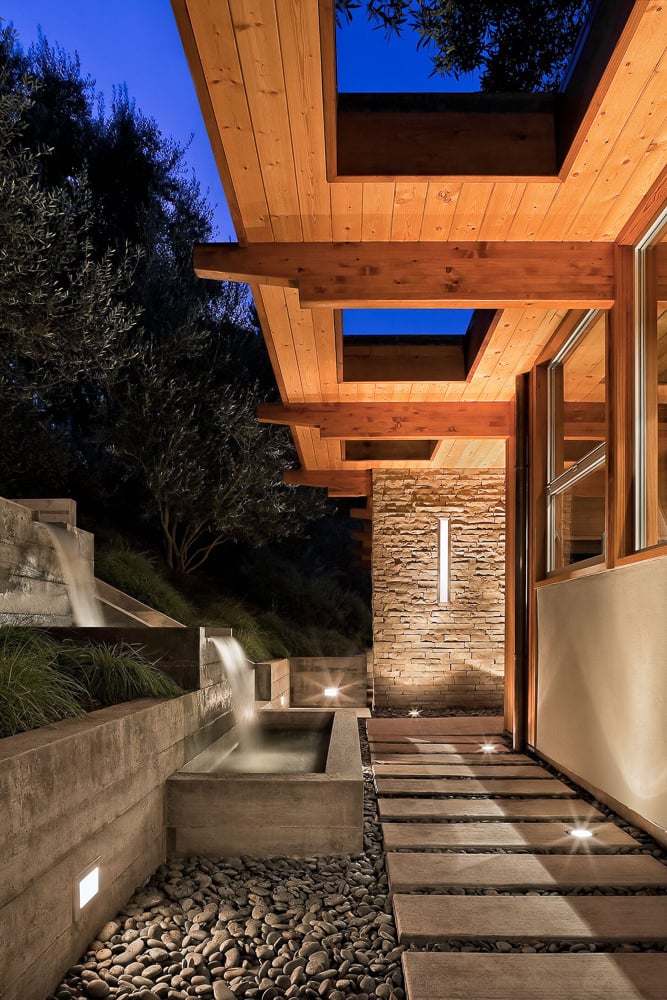 Night view of a mid-century modern entry path with wood beams, skylights, and a serene water feature.