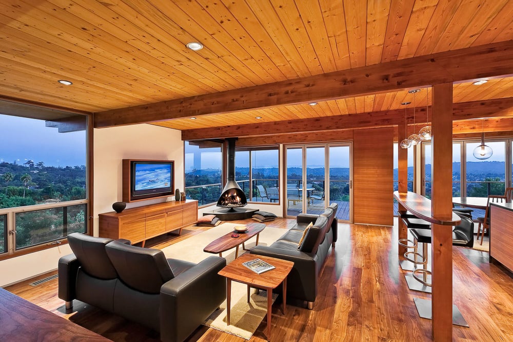 Mid-century modern living room with suspended fireplace, wood finishes, and full-height glazing connecting to a wraparound deck.