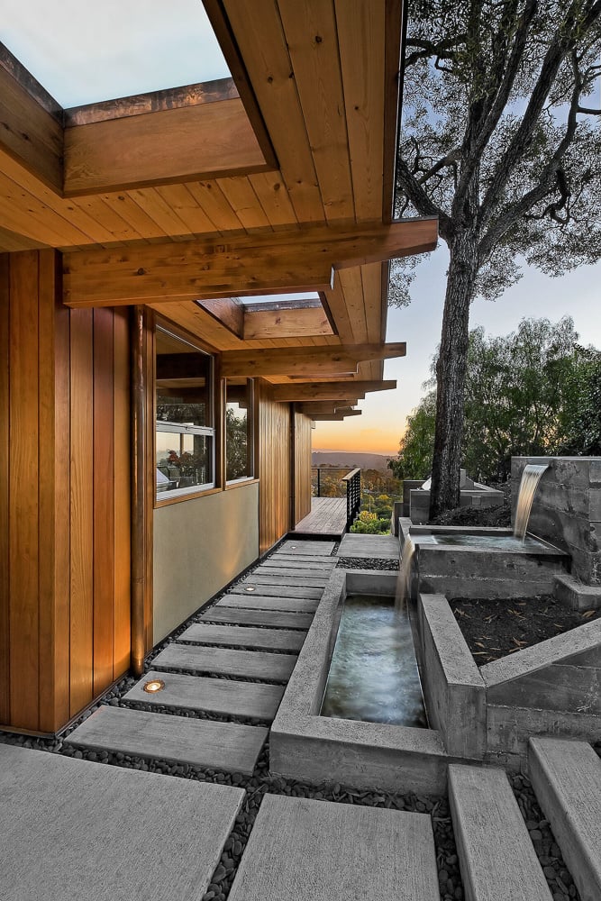 Exterior walkway of a mid-century modern home featuring exposed wood beams, skylights, and a minimalist concrete water feature.