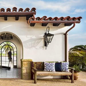 This Santa Barbara influenced Spanish style house, features a wooden bench with decorative pillows in front of a Neoclassical Spanish residential building with an entry arch, wrought iron light fixture, and copper downspout