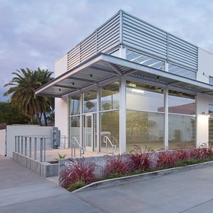 Modern commercial building exterior surrounded by board formed concrete walls featuring expansive aluminum framed storefront windows and shaded by a galvanized steel overhang and detailing