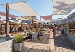 Perspective view of a bustling outdoor courtyard built with repurposed shipping container shops, featuring wooden decking, grey and pink shade sails, string lights, and patrons browsing, dining, and walking.