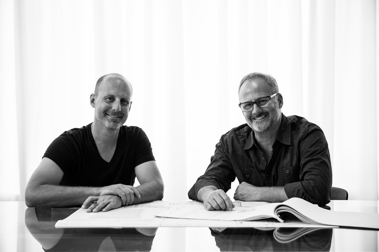Architects Josh Blumer and Clay Aurell pose for a portrait sititng at a table together with a set of drawings in front of them,