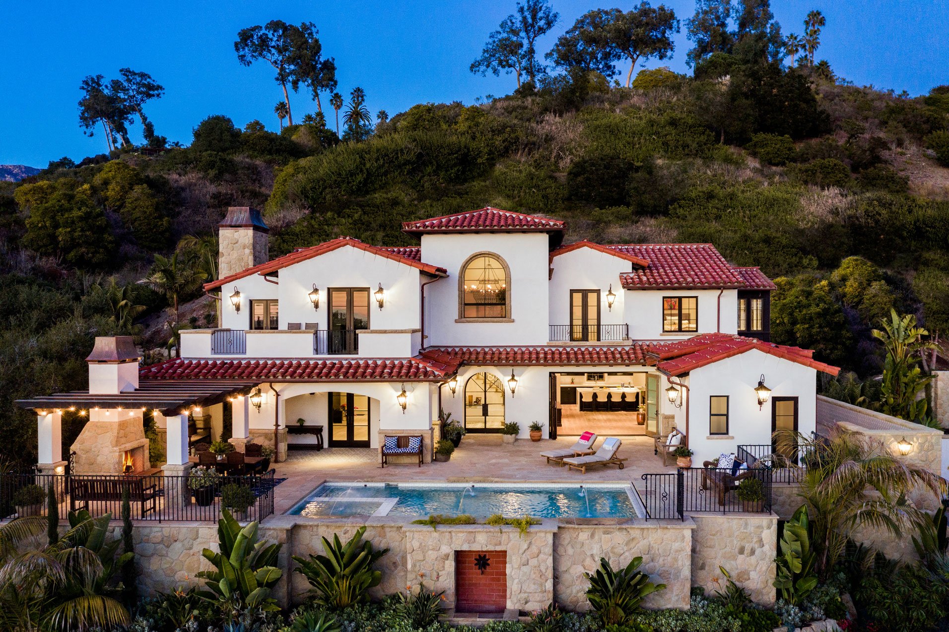 This hillside Spanish Colonial Revival home showcases traditional architecture with red clay tile roofs, white stucco walls, and a modern pool courtyard.