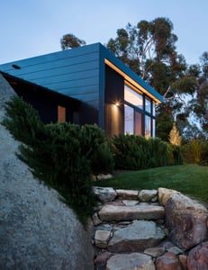 Looking up a rock pathway through beautiful landscaped property to a stunning contemporary residence clad in black metal paneling featuring expansive glass windows and a cozy interior